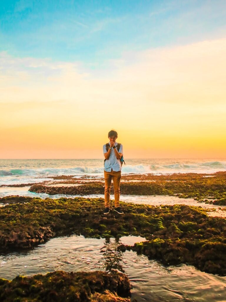 A man stands on mossy rocks by the ocean, praying during a vibrant sunset, creating a serene and introspective scene.